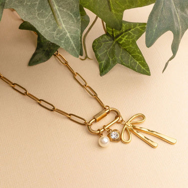 Gold necklace with bow and pearl charm on a beige background with green leaves.