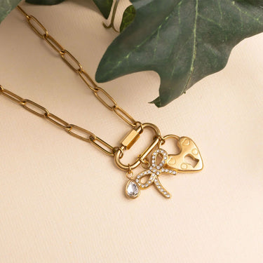Gold necklace with heart and bow-shaped charms on a beige background with green leaves.