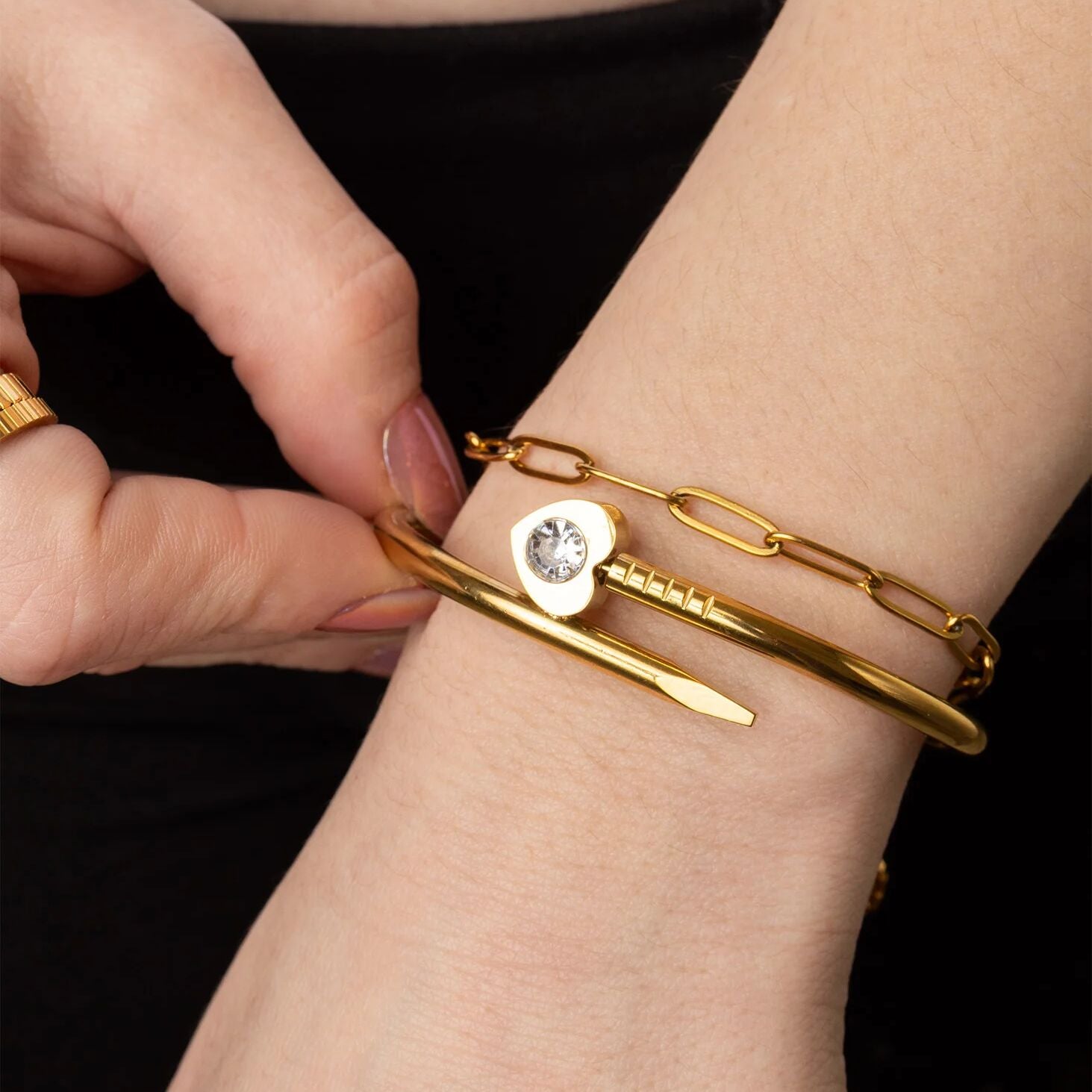 Gold bracelet with a heart-shaped charm being adjusted on a wrist against a black background