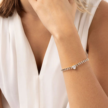 Silver bracelet with a heart-shaped charm on a person's wrist, wearing a white top.
