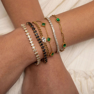 Multiple gold and silver bracelets with green gemstones on a wrist against a white fabric background