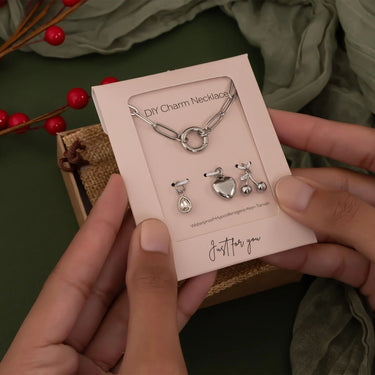 Hand holding a DIY charm necklace kit with charms on a green background