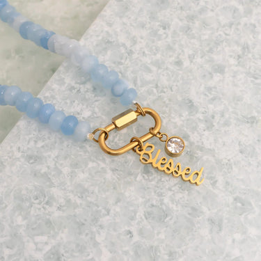 Necklace with blue beads and gold clasp with 'Blessed' charm on a textured surface