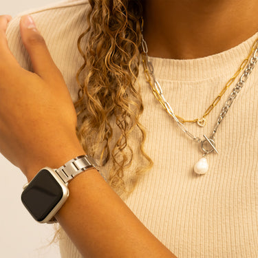 Person wearing a silver smartwatch and multiple necklaces with a neutral background