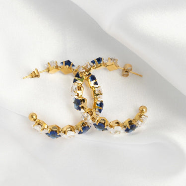 Gold hoop earrings with blue and clear gemstones on a white cloth background