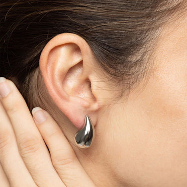 Close-up of an ear wearing a silver teardrop earring with a hand touching the earlobe.
