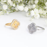 Two Floral Motif Rings, one gold and one silver, on a white surface with white flowers in the background.