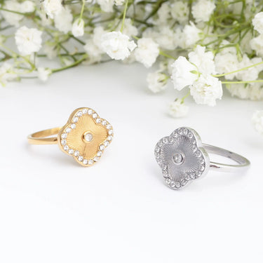 Two Floral Motif Rings, one gold and one silver, on a white surface with white flowers in the background.