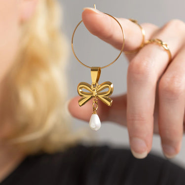 Gold hoop earring with bow and pearl pendant held by a hand against a neutral background