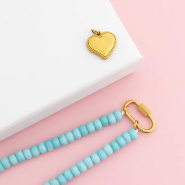 Gold heart-shaped pendant on a turquoise beaded necklace against a pink background