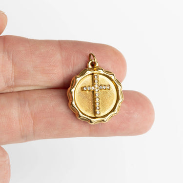 Gold pendant with a cross design held between fingers on a white background