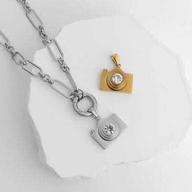 Two camera-shaped pendants, one gold and one silver, on a white background.
