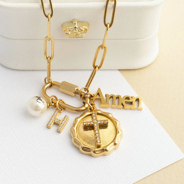 Gold necklace with charms including a cross, 'Amen', and 'H' on a white surface.