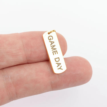 Gold pendant with 'GAME DAY' engraving held between fingers on a white background