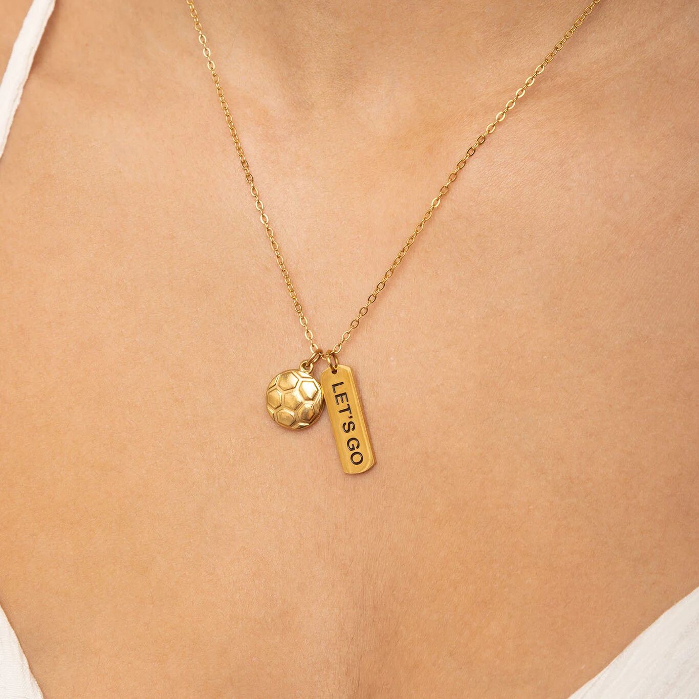 Gold necklace with a small round pendant and 'LET'S GO' tag on a person's neck.