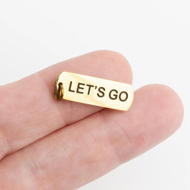 Gold charm with 'LET'S GO' text held between fingers on a white background