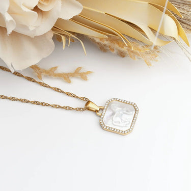 Gold necklace with a clear pendant on a white surface with flowers in the background