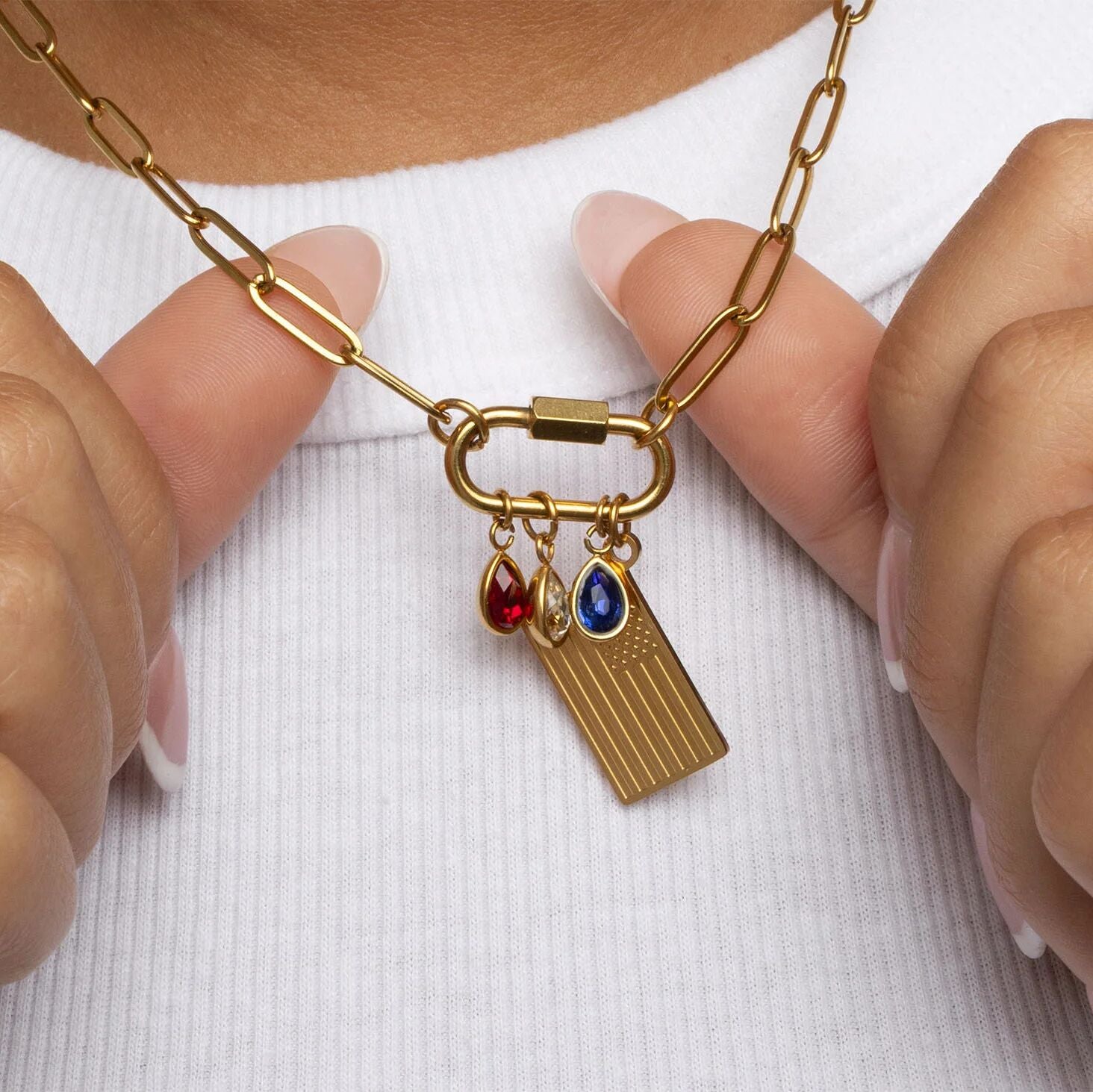 Gold necklace with Flag Charm and red, clear and blue teardrop birthstone charms held by hands against a white background