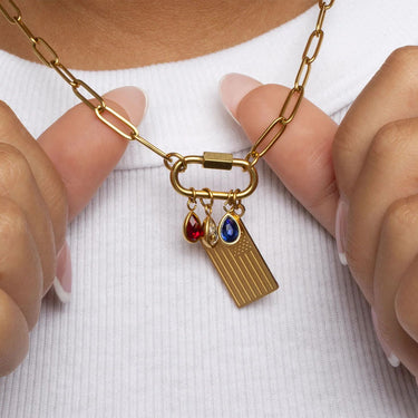Gold necklace with Flag Charm and red, clear and blue teardrop birthstone charms held by hands against a white background
