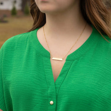 Person wearing a gold necklace with a bar pendant against a blurred outdoor background