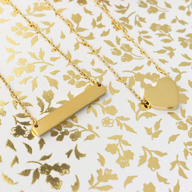Two gold necklaces, one bar-shaped and one heart-shaped, on a gold floral patterned background.