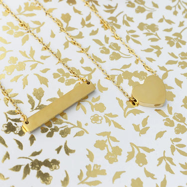 Two gold necklaces, one with a bar pendant and the other with a heart locket, on a floral patterned background.