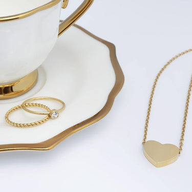 Gold necklace with heart pendant on a white plate with gold rim, next to two gold rings.