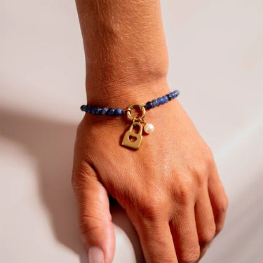 Hand wearing a blue beaded bracelet with a gold charm and pearl on a neutral background