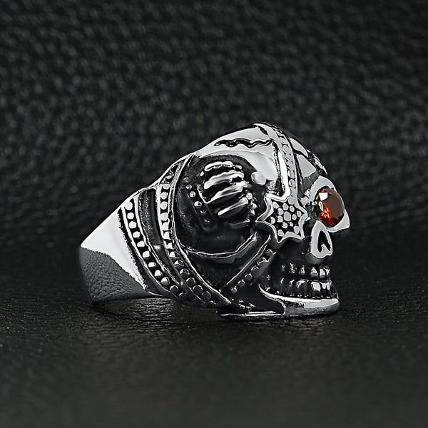 Stainless steel skull with red Cubic Zirconia eye and eyepatch ring angled on a black leather background.
