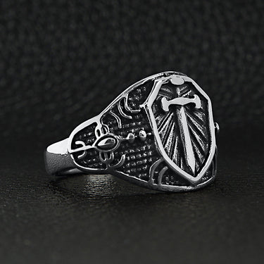 Stainless steel medieval sword and shield ring angled on a black leather background.
