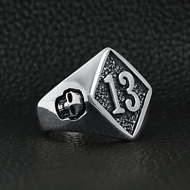 Stainless steel "13" and skulls ring angled on a black leather background.