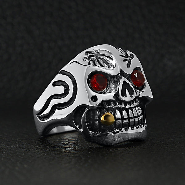 Stainless steel 18K gold plated bullet cigar smoking red Cubic Zirconia eyed skull ring angled on a black leather background.
