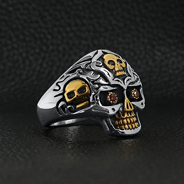 Stainless steel 18K gold PVD Coated red Cubic Zirconia eyed filigree skull ring angled on a black leather background.