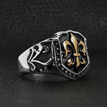 Stainless steel 18K gold PVD Coated Fleur De Lis shield ring angled on a black leather background.