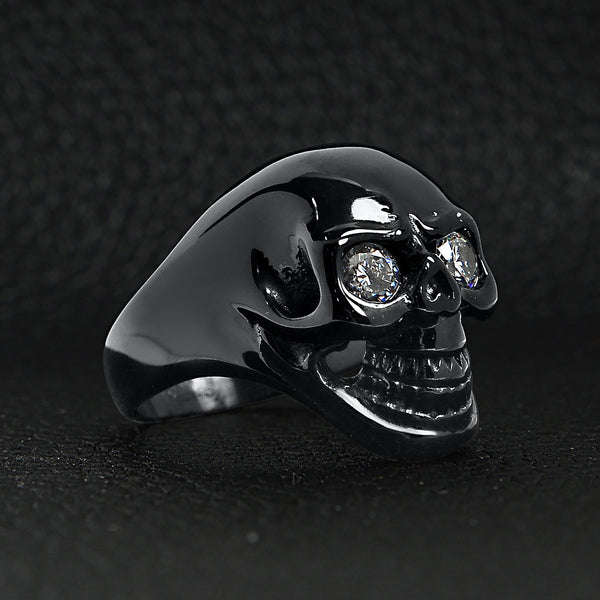 Stainless steel clear Cubic Zirconia eyed black skull ring angled on a black leather background.