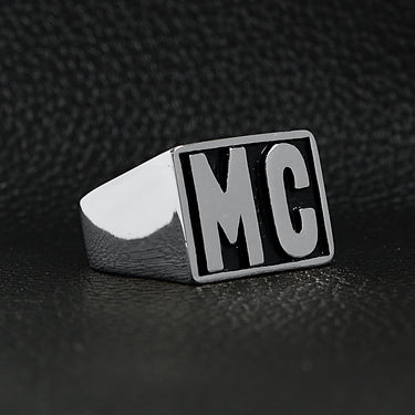 Stainless steel motorcycle club "MC" insignia signet ring angled on a black leather background.