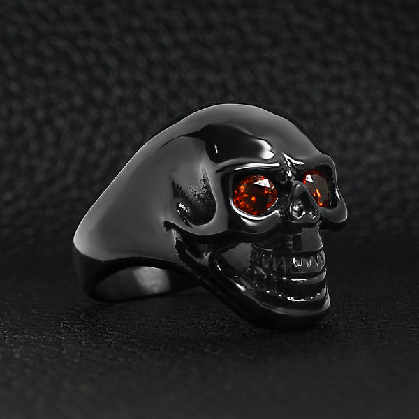 Stainless steel red Cubic Zirconia eyed black skull ring angled on a black leather background.
