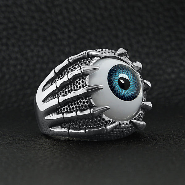 Stainless steel skeleton hands holding blue eyeball ring angled on a black leather background.